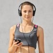 Isolated shot of young sportswoman has healthy athleteic body, listens music with headphones, holds smart phone, looks directly at camera, has cardio training, poses against grey concrete wall